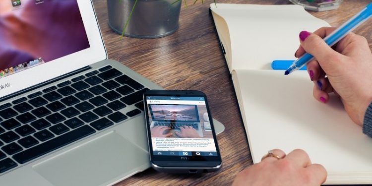woman-sitting-at-table-writing-in-notepad-with-laptop-and-mobile-phone-on-wooden-table